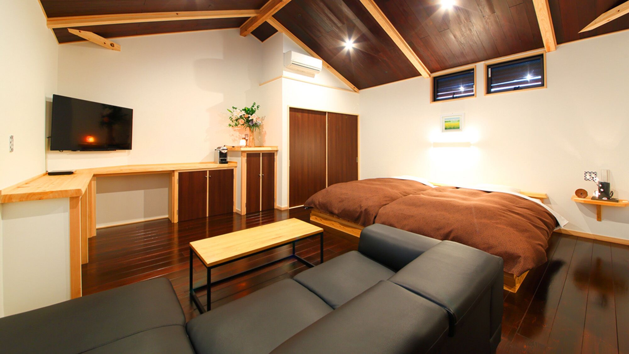 A compact room with a bed, a sofa, a small table, a TV, and a kitchenette area.