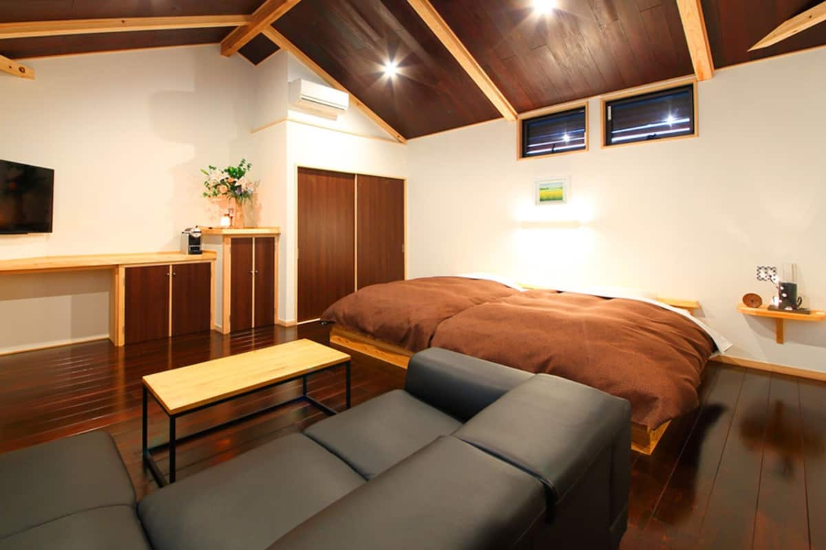 A compact room with a bed, a sofa, a small table, a TV, and a kitchenette area.