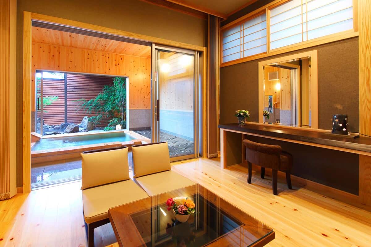 A room with a glass table, a sofa, a small desk, and a view of a garden and outdoor area.