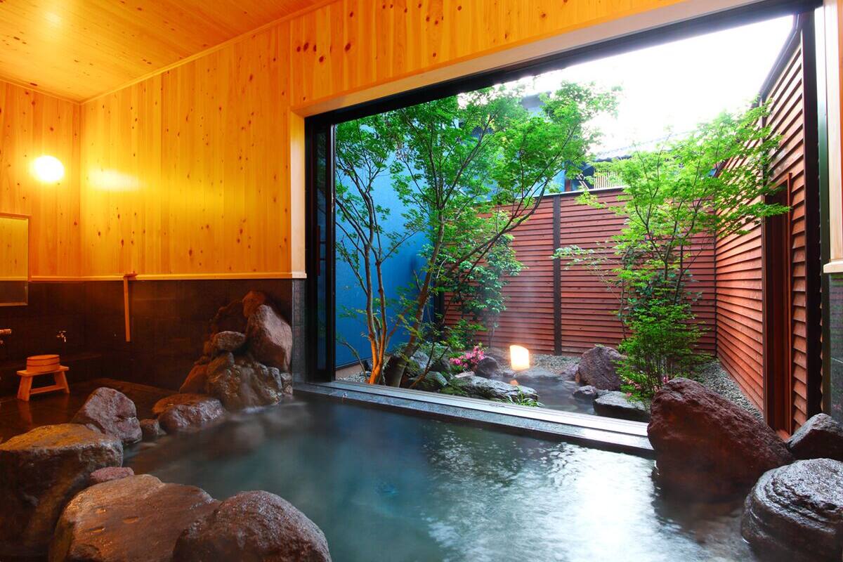 A traditional Japanese onsen with a wooden interior, a view of greenery outside, and a stone bath area.