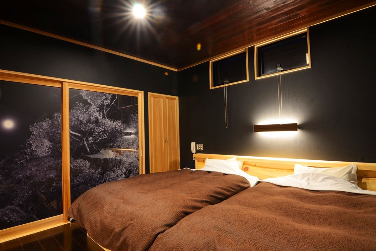 A bedroom with a large bed, a nightstand, a wall-mounted lamp, and a window with a view of trees.