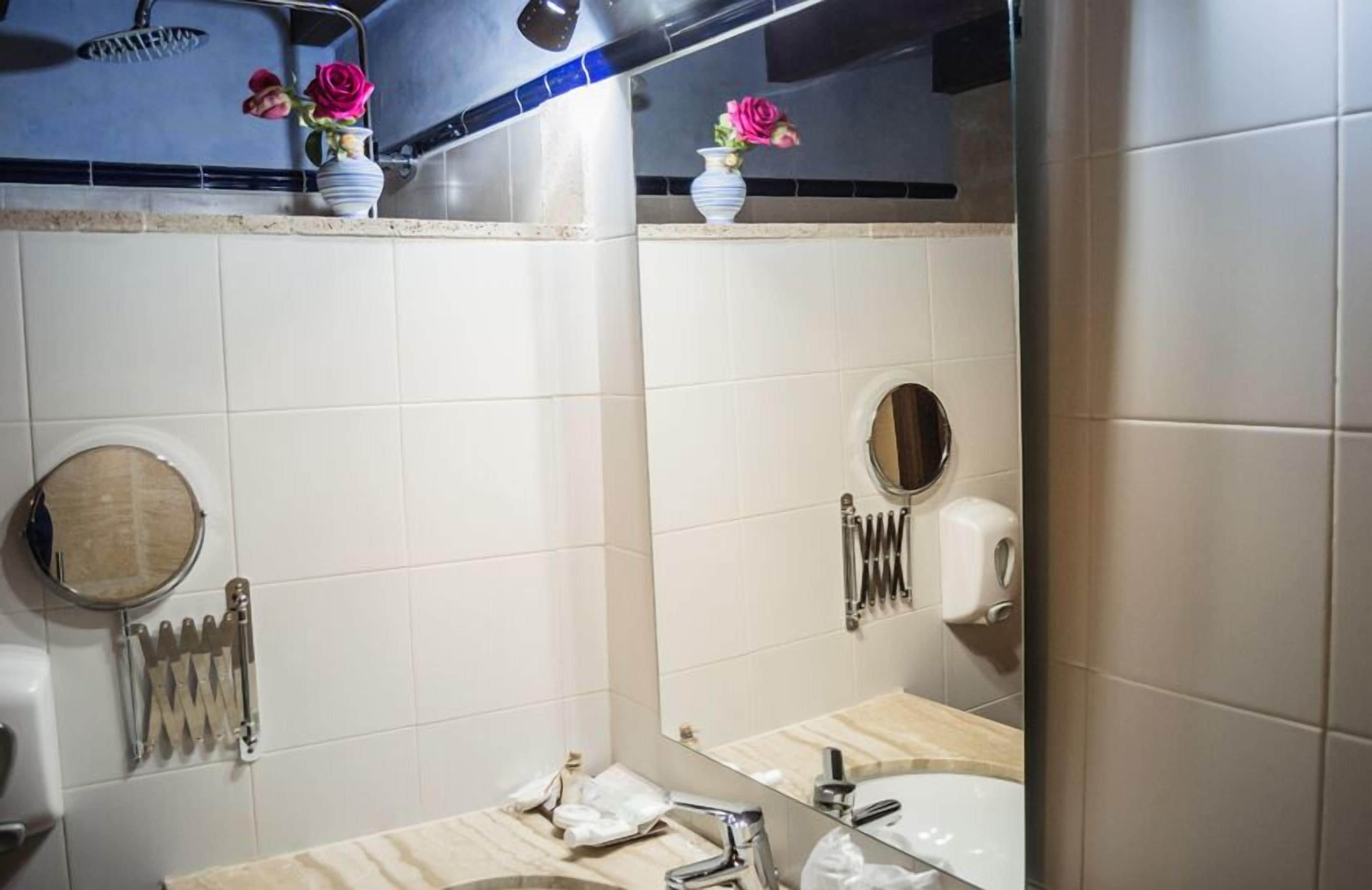 Une salle de bain avec des carreaux blancs, un lavabo, un miroir, un porte-serviettes et une pomme de douche.