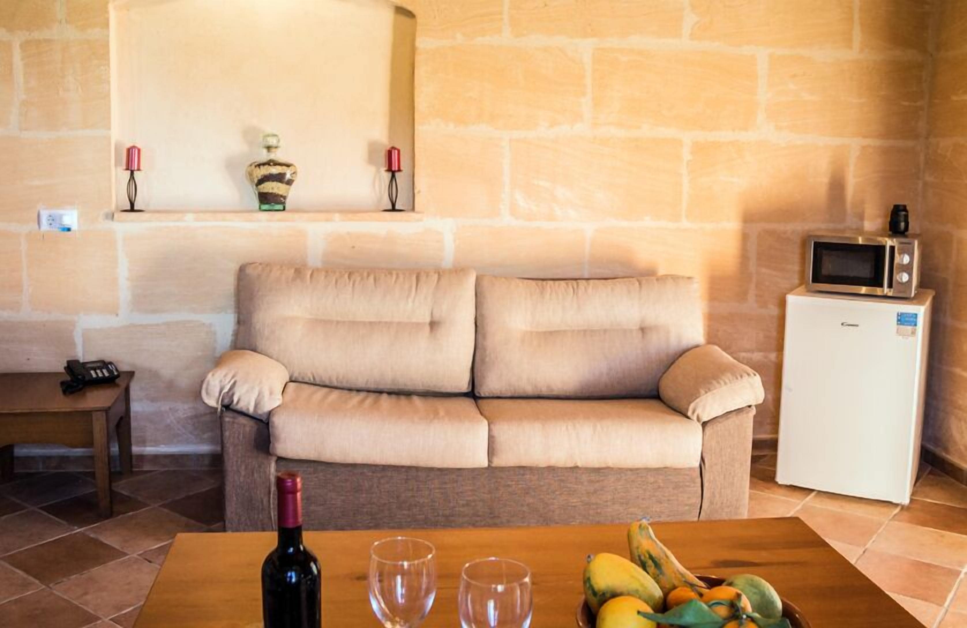 Un salón acogedor con un sofá beige, una mesa de centro de madera con una botella y vasos, un televisor pequeño en un mueble blanco y una pared de piedra.