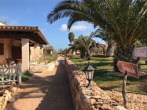 Un chemin menant à un bâtiment doté d’un toit en tuiles, de palmiers et de murs en pierre.