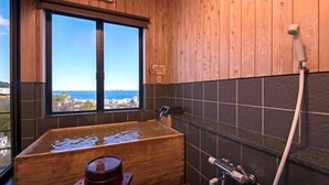 A wooden cabin bathroom with a large bathtub, a window with a view of the sea, and a shower area. - Ayunokaze (Ito)