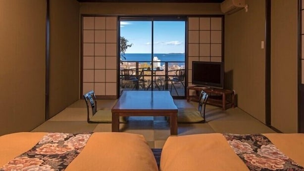 A traditional Japanese room with tatami flooring, a low table, two chairs, a television on a wooden stand, and a view of the sea through shoji screens. - Ayunokaze (Ito)