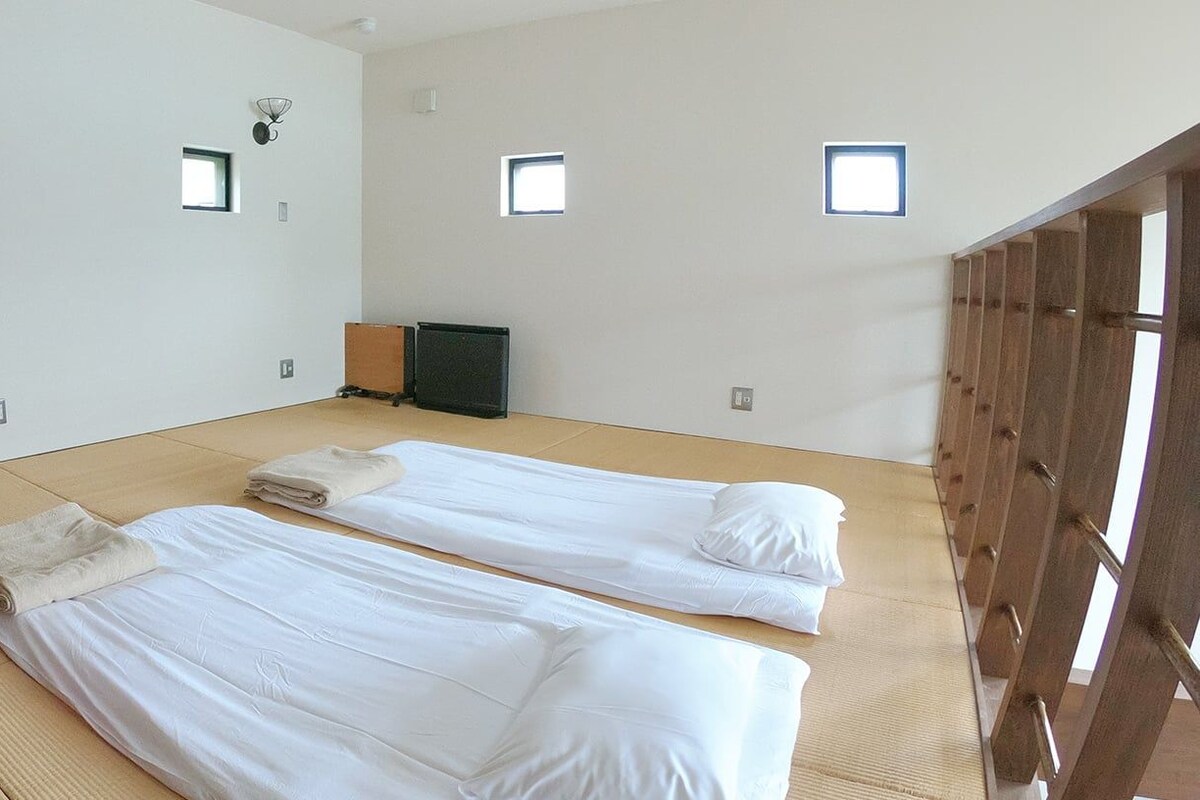 Una habitación con dos camas, esteras de tatami, ventanas pequeñas y una puerta de madera.