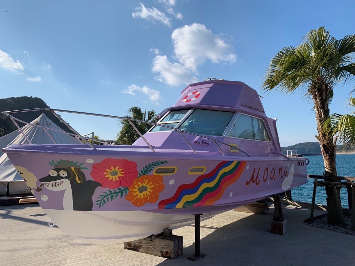 A boat with colorful floral and abstract designs, moored at a beach with palm trees and a clear sky.