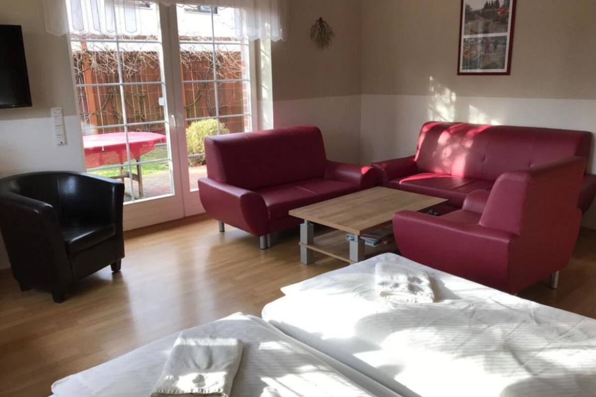 A room with a red sofa, a black chair, a wooden table, and a bed with white bedding.