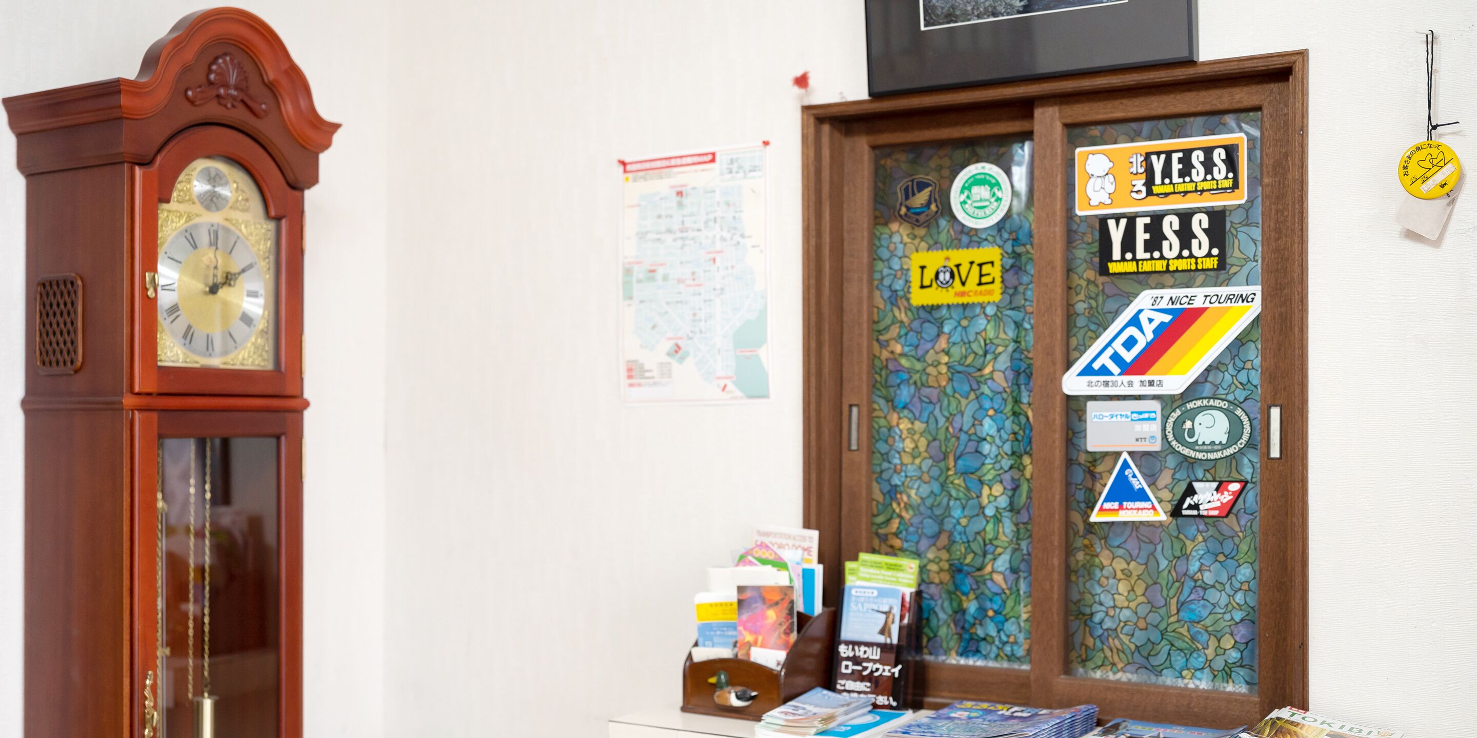 Una habitación con un reloj de pie de madera, una puerta decorada con pegatinas y una pequeña mesa con libros y revistas.