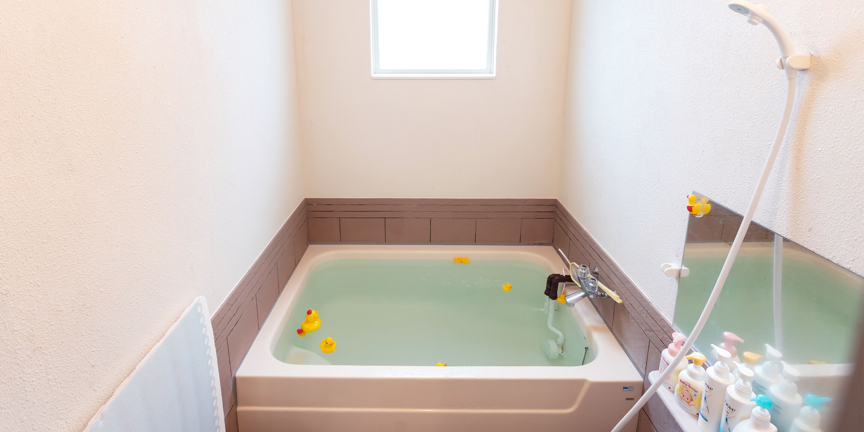 A bathroom with a bathtub containing rubber ducks, a window, and toiletries on a shelf.