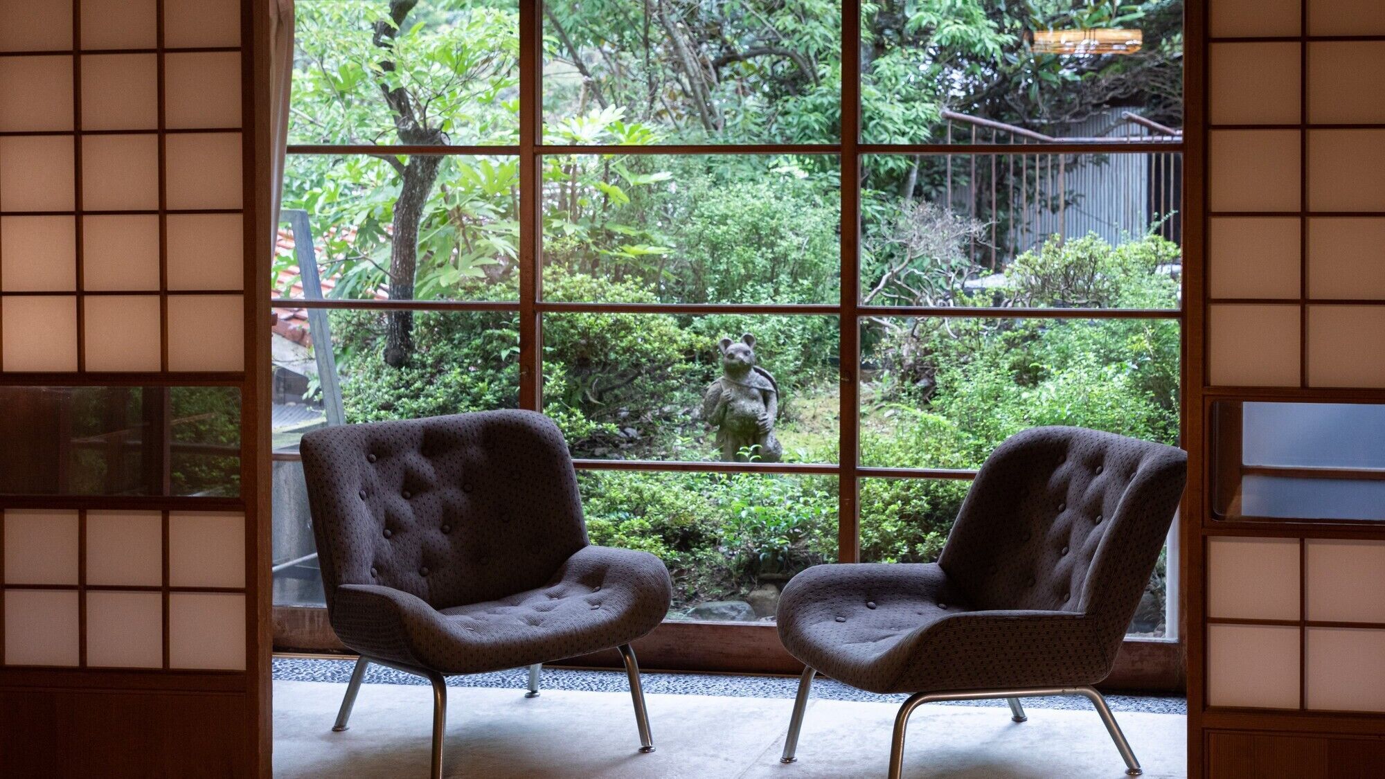 A room with two upholstered chairs, a large window with a grid pattern, and a view of a garden with a statue.