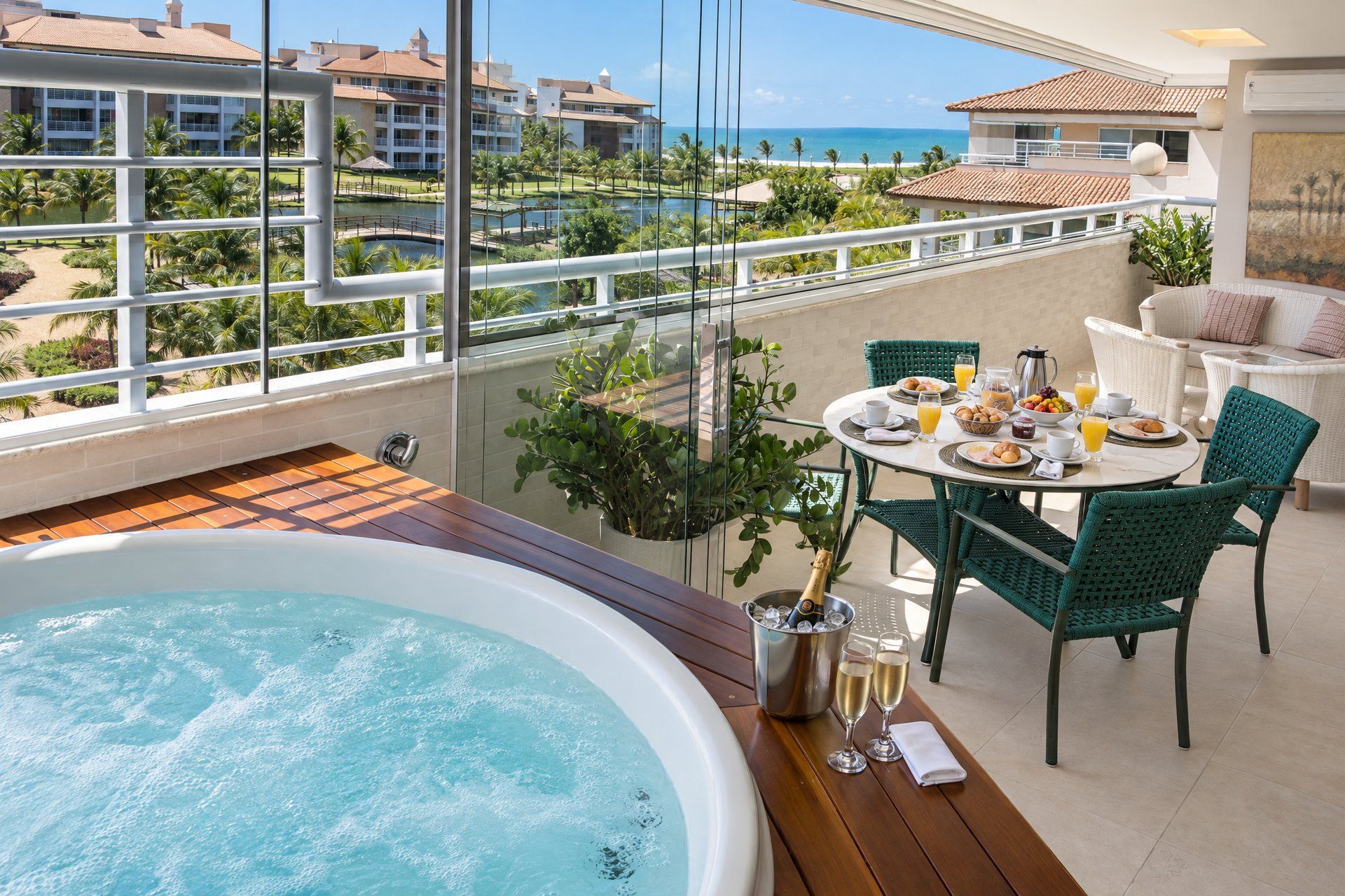 A balcony with a hot tub, dining table, chairs, and a view of the ocean and resort buildings.