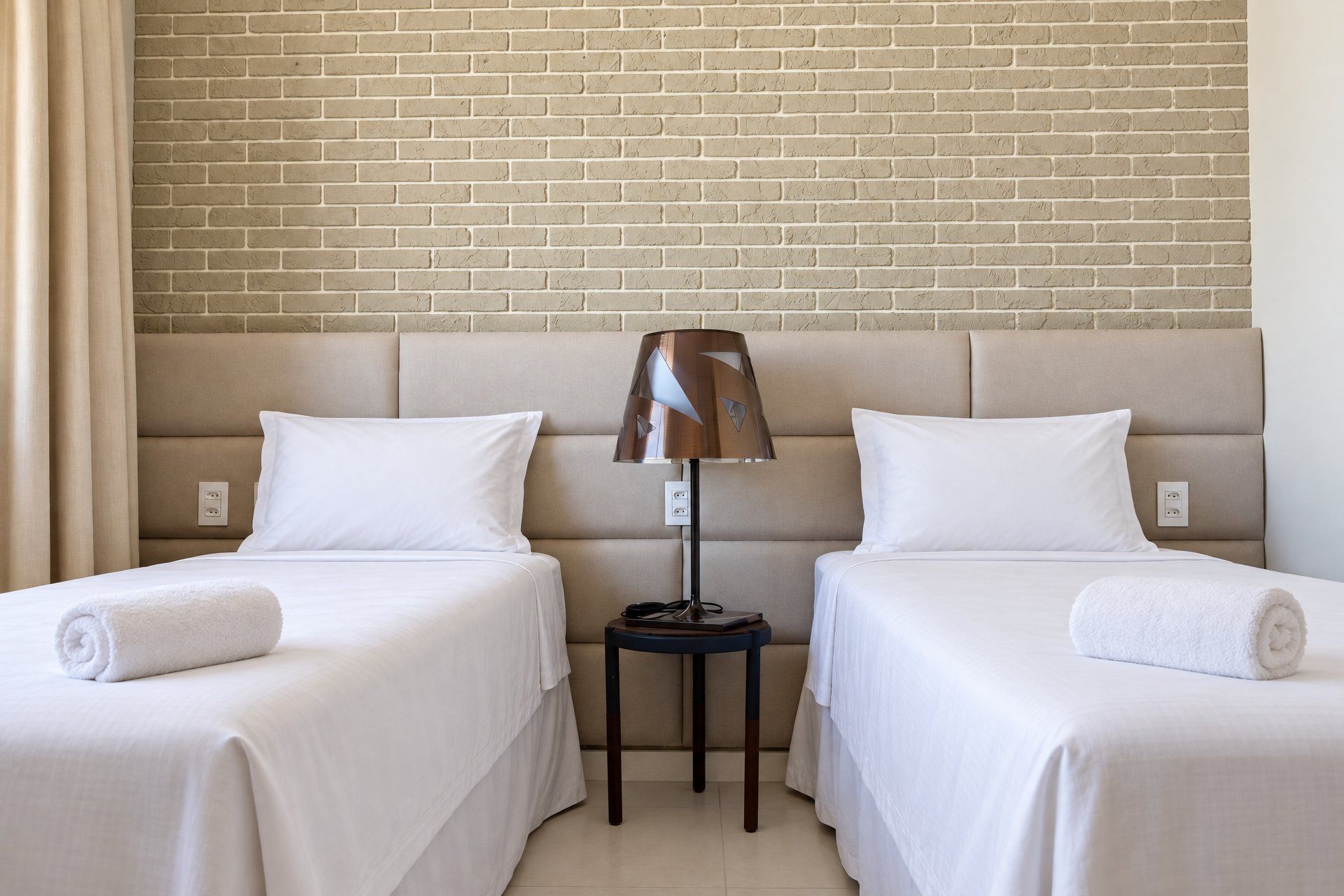Una habitación de hotel con dos camas individuales, una mesita de noche con lámpara y una pared de ladrillo.