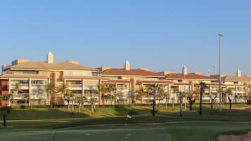 Um campo de golfe com bandeira, palmeiras e um hotel com várias varandas e telhado vermelho.
