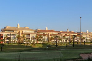 A golf course with a flag, palm trees, and a hotel with multiple balconies and a red roof. - Penthouse Apartment with Hot Tub near Beach (Aquiraz)