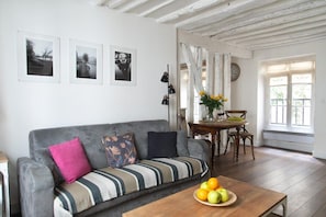 Uma sala de estar com um sofá cinzento, uma mesa de centro de madeira com frutas, uma mesa de jantar com cadeiras e uma janela com cortinas.