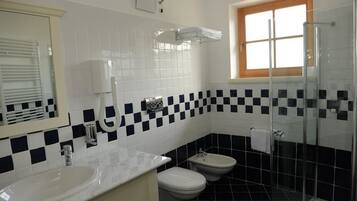 A bathroom with a white sink, two toilets, a bidet, a glass shower enclosure, and a window with blinds.