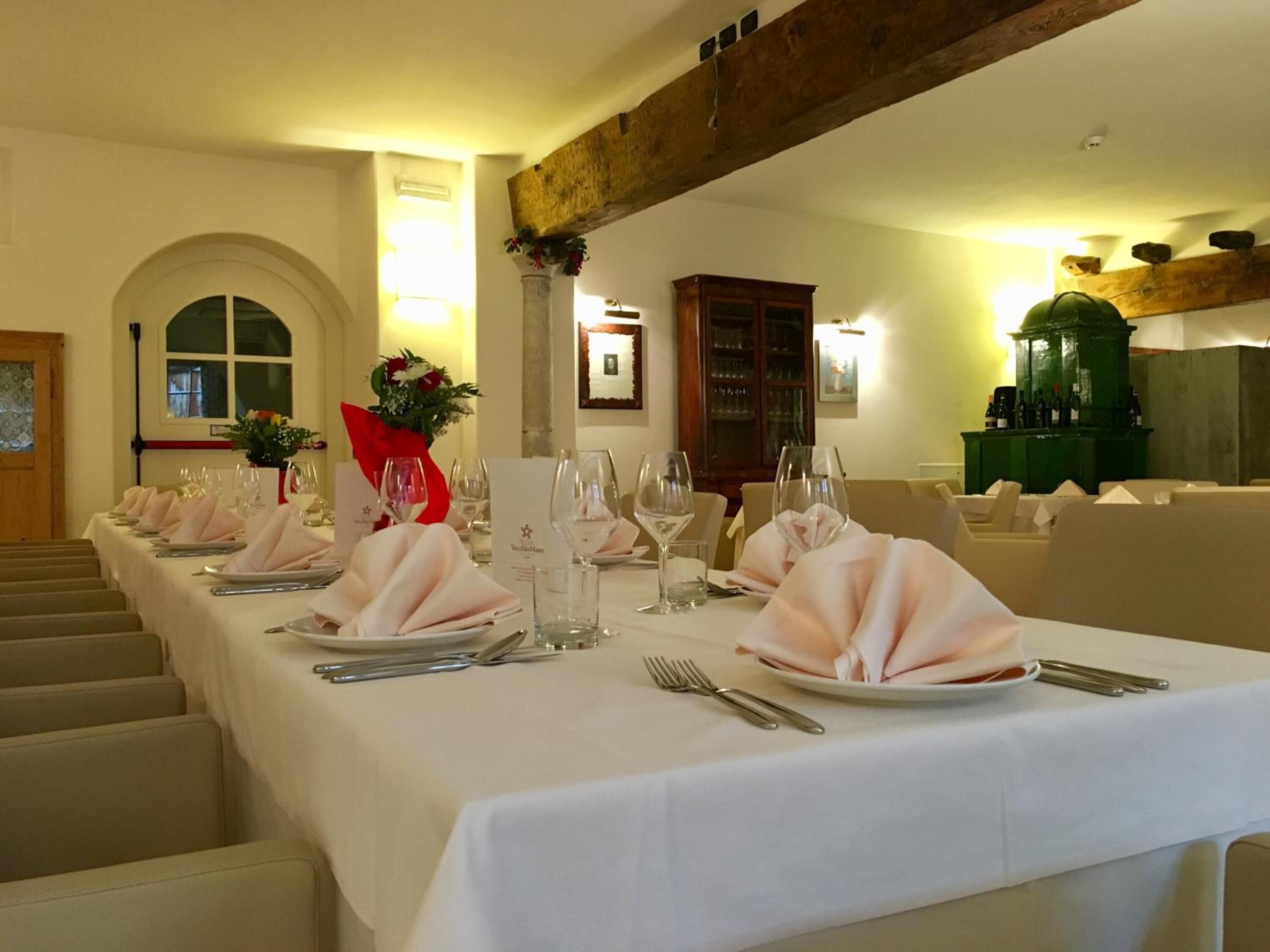Ein langer Esstisch, gedeckt mit weißen Tischdecken, rosa Servietten und Besteck, unter einer Holzbalkendecke mit einem grünen Schrank im Hintergrund.
