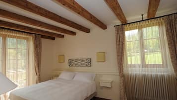 Une chambre à coucher avec un lit, des poutres en bois, une table de chevet, une lampe fixée au mur et une fenêtre avec des rideaux.