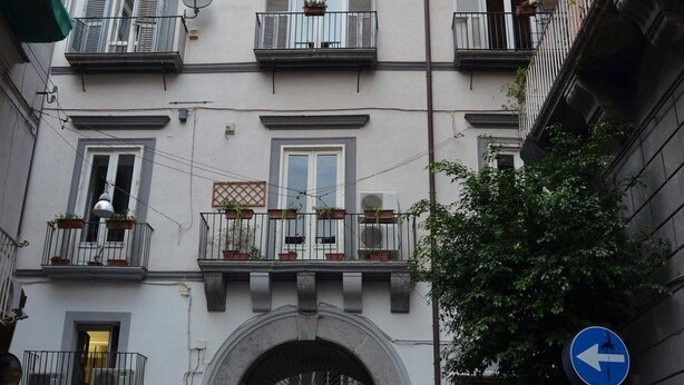 Un edificio de varios pisos con balcones, una entrada con arco central y un mercado de flores en el exterior.