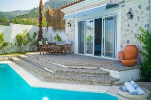 A poolside area with a stone wall, a table with chairs, and a thatched roof. - ALYA COUNTRY (Marmaris)