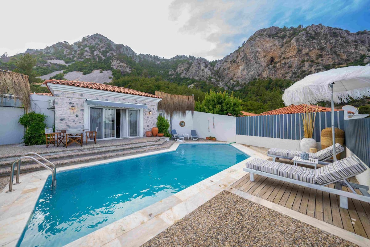 Une piscine entourée de chaises longues et d’une table, le tout bordé d’une terrasse en bois et avec une chaîne de montagnes en toile de fond.