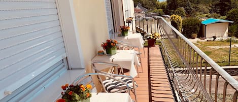 A balcony with white tables and chairs, potted plants, and a view of a forested area.