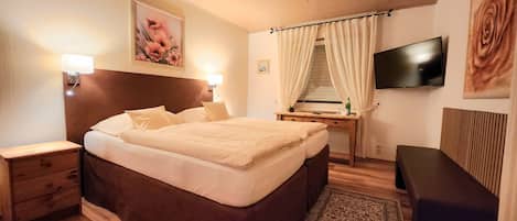 Habitación de hotel con cama de madera, mesitas de noche, un escritorio con silla, televisor montado en la pared y un ventanal con cortinas.