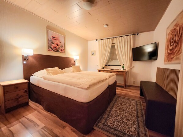 Une chambre d’hôtel avec un lit en bois, des tables de chevet, un bureau avec une chaise, un téléviseur fixé au mur et une grande fenêtre avec des rideaux.