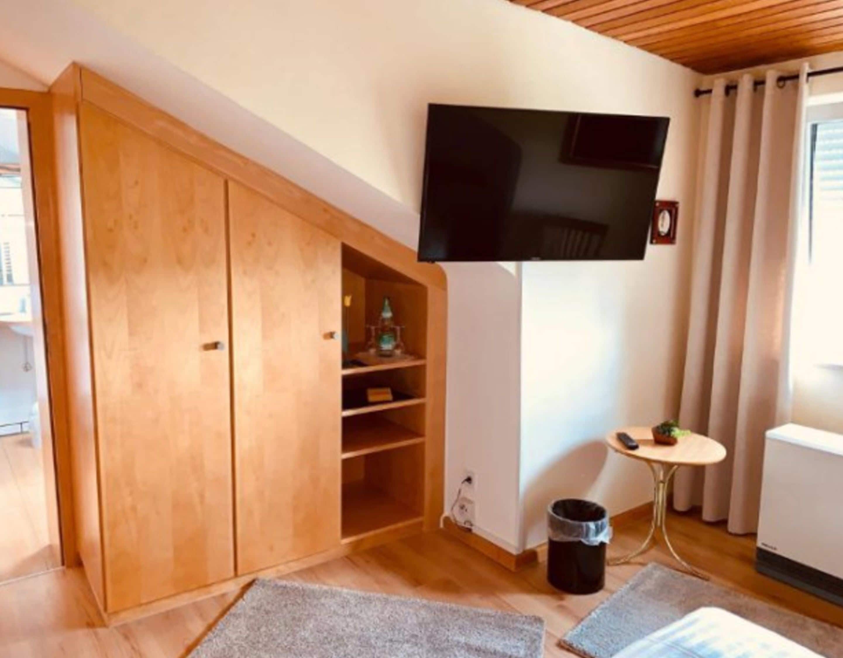 A room with a wooden wardrobe, a mounted TV, a small table with a chair, and a radiator.