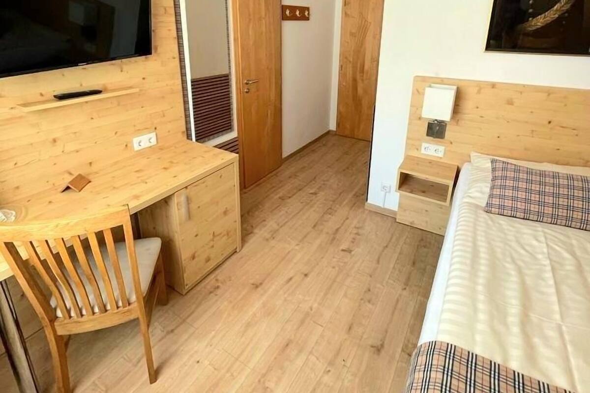 Een compacte hotelkamer met een houten bureau, een bed met een geruit dekbedovertrek, een wandgemonteerde televisie en een ingelijst schilderij.