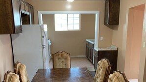 Une salle à manger avec une grande table en bois et six chaises rembourrées. On trouve un réfrigérateur et un four à micro-ondes sur le côté gauche, et une fenêtre qui laisse entrer la lumière naturelle.