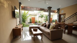 A spacious living room with a sofa, coffee table, and a dining area with a wooden table and chairs. There is a ceiling fan, a TV mounted on the wall, and a staircase leading to an upper level. - Kubu Willis (Legian)