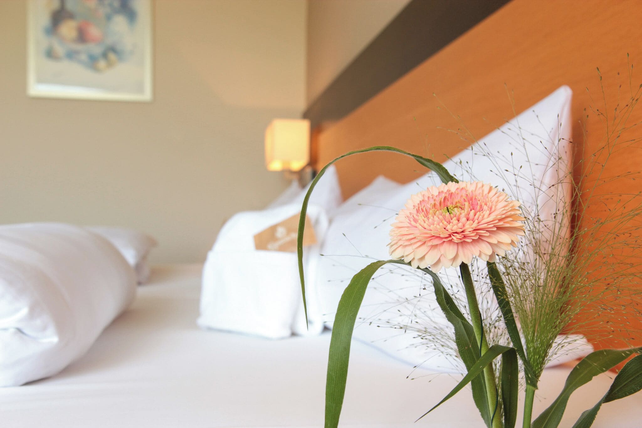 Un lit bien fait, avec du linge de lit blanc, une seule fleur rose et un élément décoratif mural.