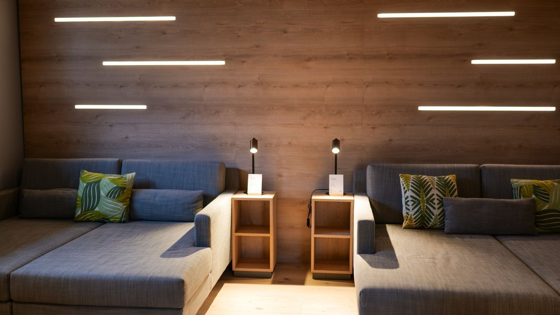 A modern living room with two sofas, each with a side table and a lamp. The room features a wooden wall with horizontal light strips.