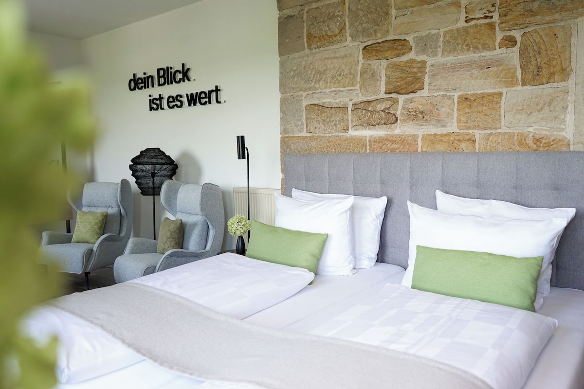 A hotel room with a stone wall, a bed with white and green bedding, a grey upholstered headboard, and a wall-mounted lamp.