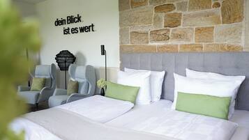 A hotel room with a stone wall, a bed with white and green bedding, a grey upholstered headboard, and a wall-mounted lamp.