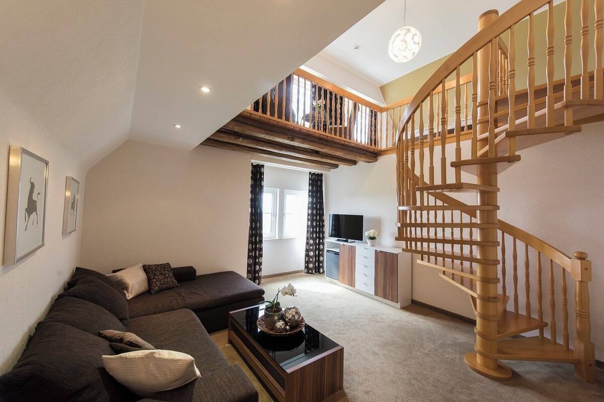 A spacious living area with a modern sofa set, a wooden staircase, and a television in the background.