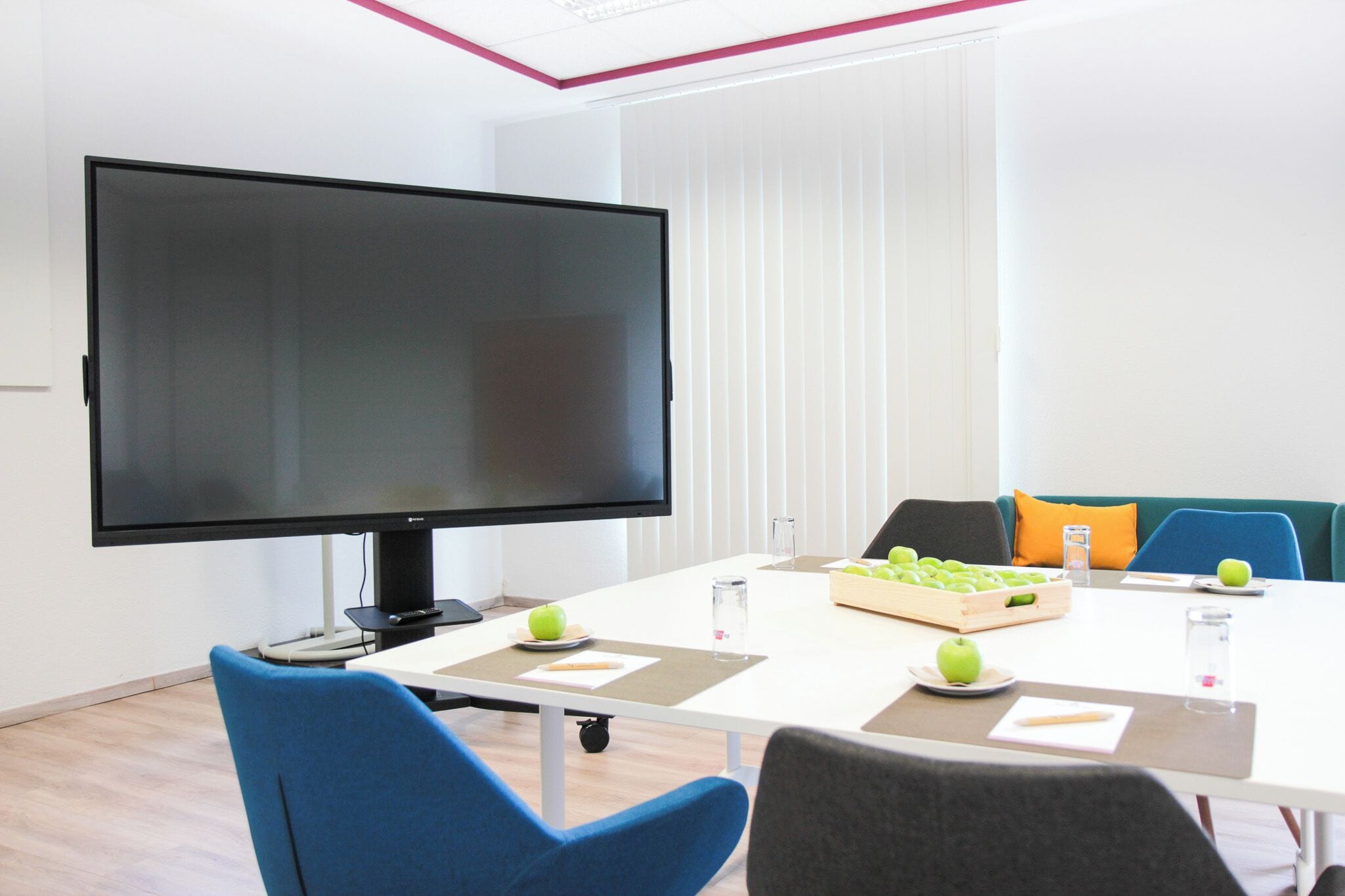 A conference room with a large flat-screen TV, a white table, and blue and gray chairs. There are apples and a glass of water on the table.
