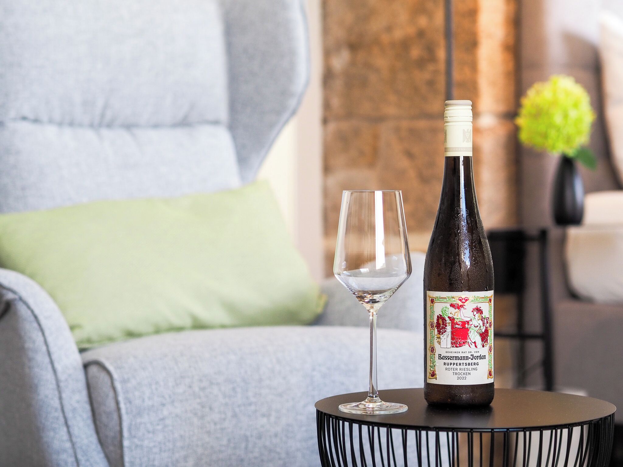 Una botella de vino y una copa vacía sobre una mesa redonda pequeña en una habitación con un sillón gris y un cojín verde.