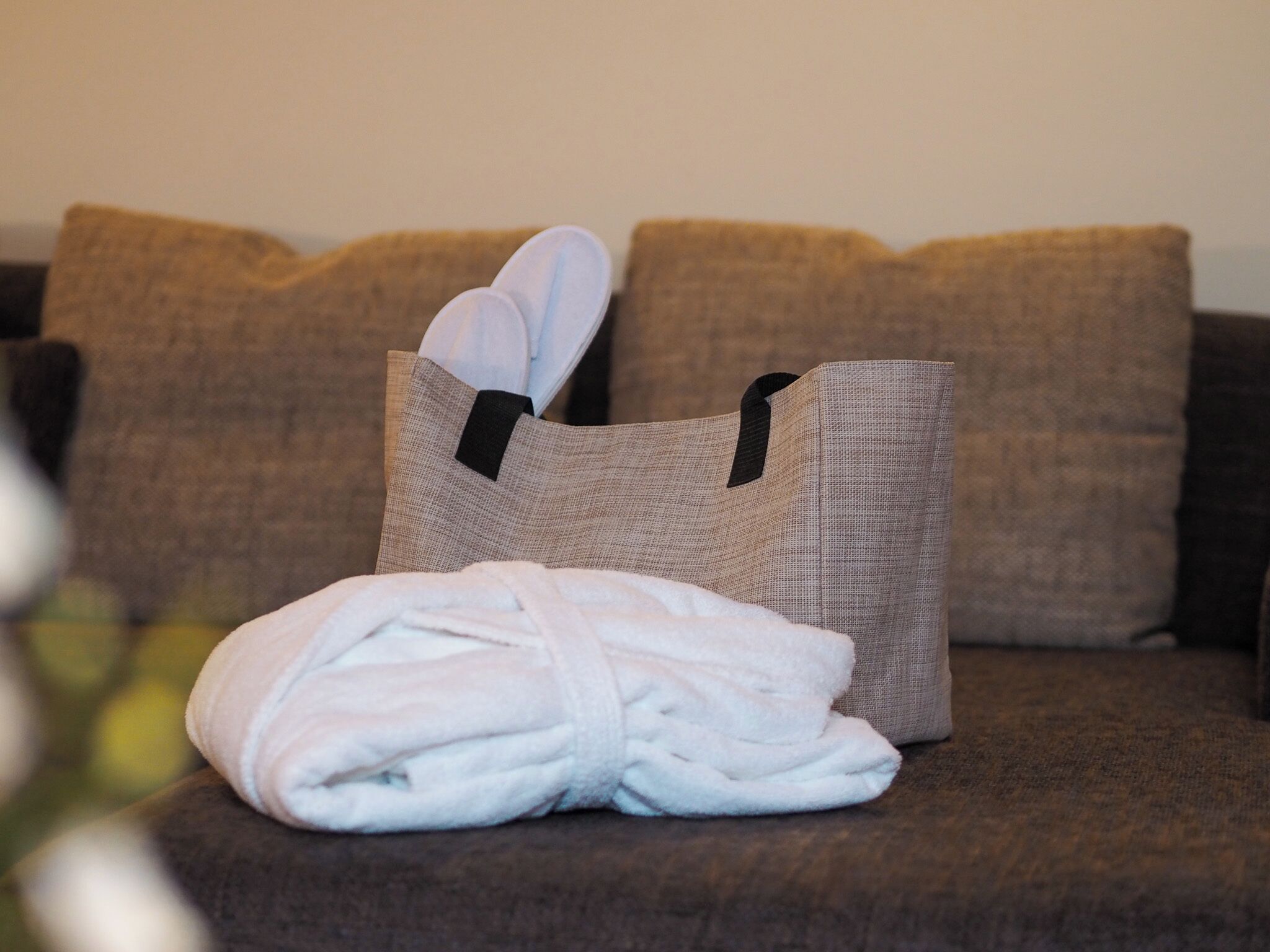 A neatly folded white bathrobe on a brown couch with a woven bag containing slippers.