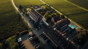 An aerial view of a vineyard with rows of grapevines, a parking lot with several cars, and a building with a sloped roof. - Leinsweiler Hof (Leinsweiler)