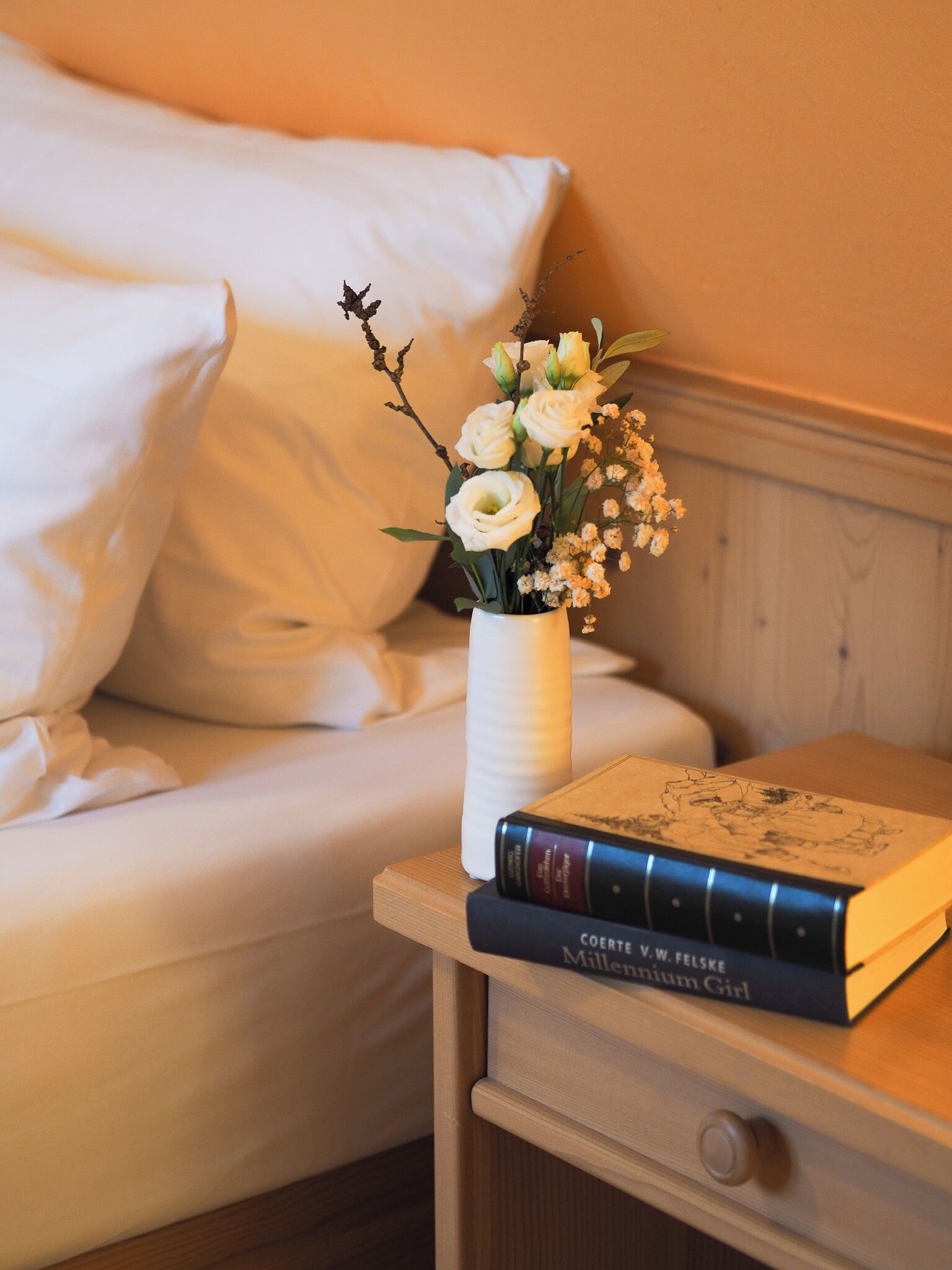 A bed with white linens, a bedside table with a book and a vase of flowers.