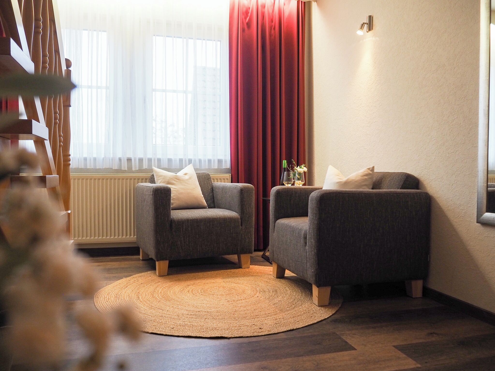 A cozy living room with two armchairs, a woven rug, a radiator, and a window with sheer curtains.