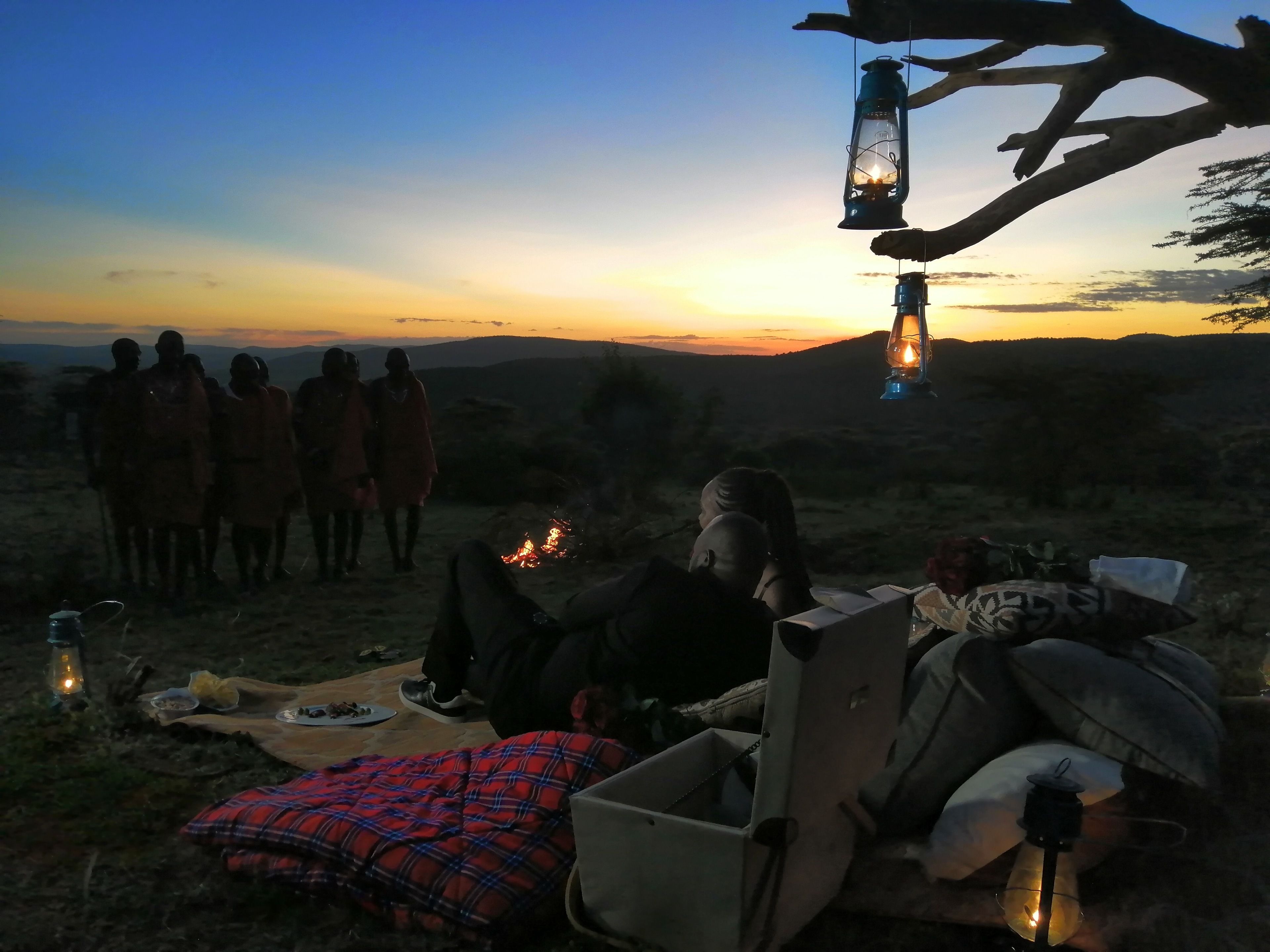 Un grupo de personas reunidas alrededor de una fogata al anochecer, con una vista despejada del atardecer al fondo.