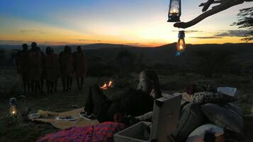 Un grupo de personas reunidas alrededor de una fogata al anochecer, con una vista despejada del atardecer al fondo.