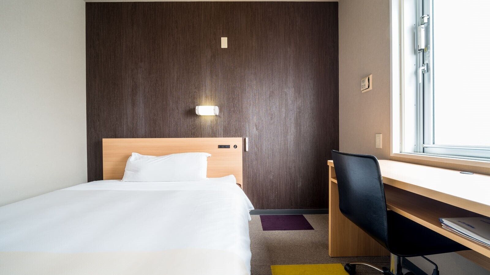 Quarto de hotel com cama, escrivaninha, cadeira, janela e luminária na parede.