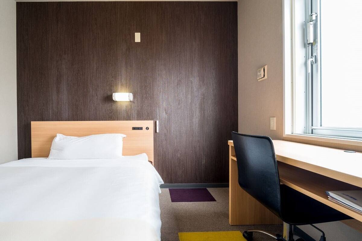 A hotel room with a bed, a desk, a chair, a window, and a wall-mounted light.