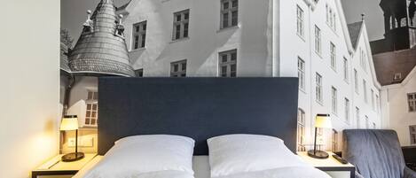 Habitación de hotel con una cama grande, dos mesitas de noche con lámparas, una silla y vistas a un edificio blanco con múltiples ventanas y una torre.